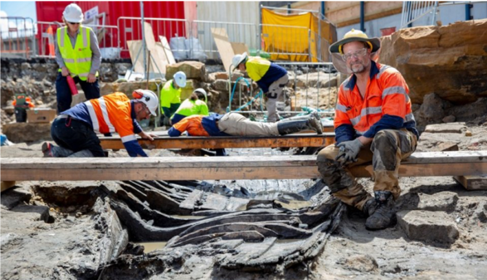 Photo of archaeologists and Karina Acton (in the background) at the dig site (9News (2018), Historic 180 year old boat uncovered in excavations for Sydney Metro station at Barangaroo, https://www.9news.com.au/2018/11/15/16/30/sydney-metro-historic-1830s-boat-uncovered-in-excavations-at-barangaroo).