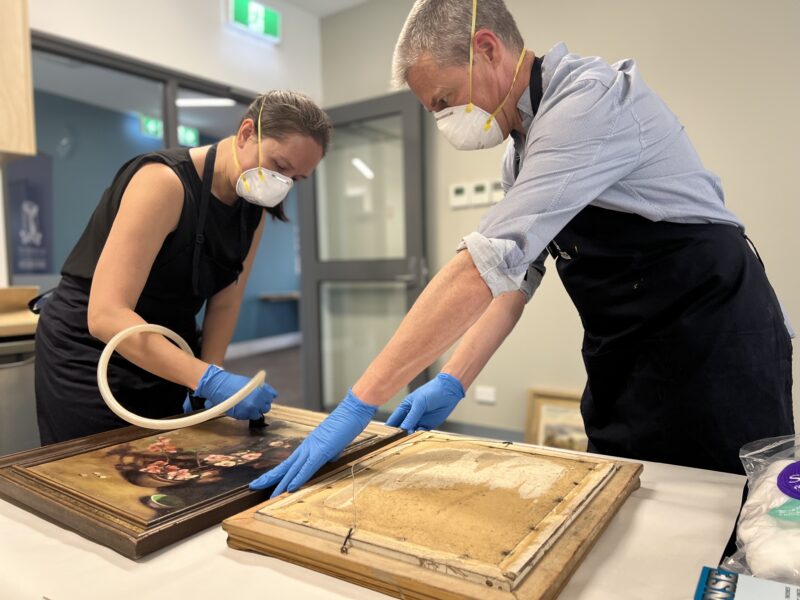 Two conservators wearing masks and gloves brush vacuuming framed works on canvas